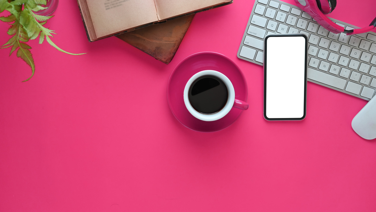 top view of a pink desk with a cup of coffee and an open book, phone and laptop on a pink background